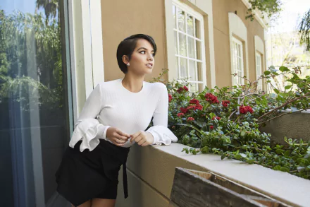 HD desktop wallpaper of a young woman with short black hair in a white blouse leaning on a balcony railing amid red flowers, gazing thoughtfully to the side.
