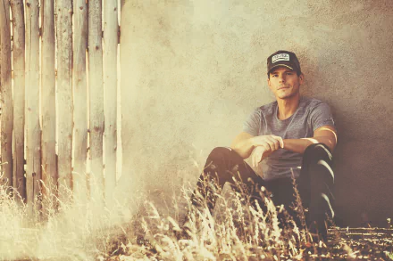 A captivating HD desktop wallpaper featuring Granger Smith seated against a textured wall, surrounded by golden grass, embodying a warm, rustic atmosphere.