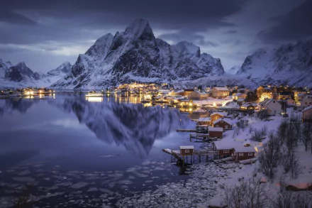 Nighttime HD desktop wallpaper of Lofoten’s snow-covered mountains reflecting in a calm lake, with village lights glowing under a cloudy sky.
