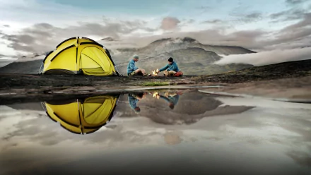 A vibrant yellow tent sits near a calm water body, reflecting the surrounding mountains. Campers enjoy a fire as clouds float overhead, capturing the essence of outdoor photography and camping.