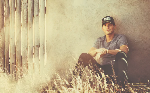 A captivating HD desktop wallpaper featuring Granger Smith seated against a textured wall, surrounded by golden grass, embodying a warm, rustic atmosphere.