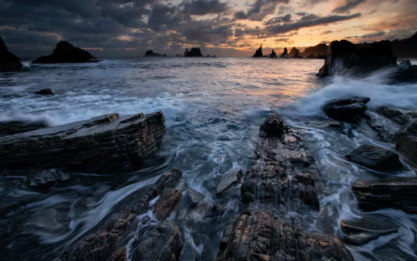 HD desktop wallpaper featuring a dramatic seascape with waves crashing against jagged rocks under a moody, cloudy sky at sunset.