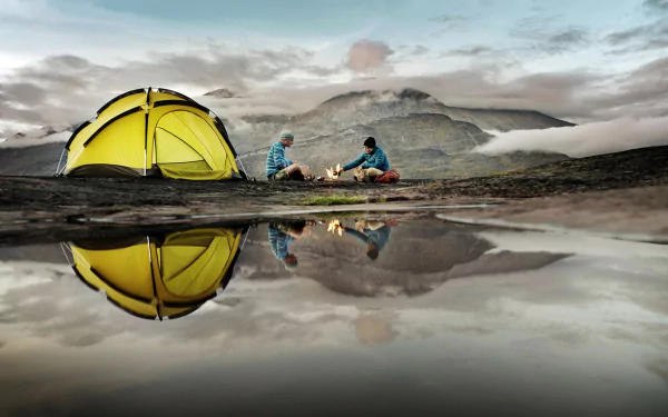 A vibrant yellow tent sits near a calm water body, reflecting the surrounding mountains. Campers enjoy a fire as clouds float overhead, capturing the essence of outdoor photography and camping.