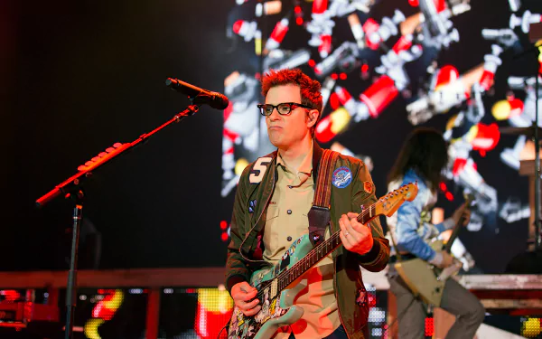 HD desktop wallpaper featuring a Weezer band member playing guitar on stage with dynamic lighting and confetti in the background.