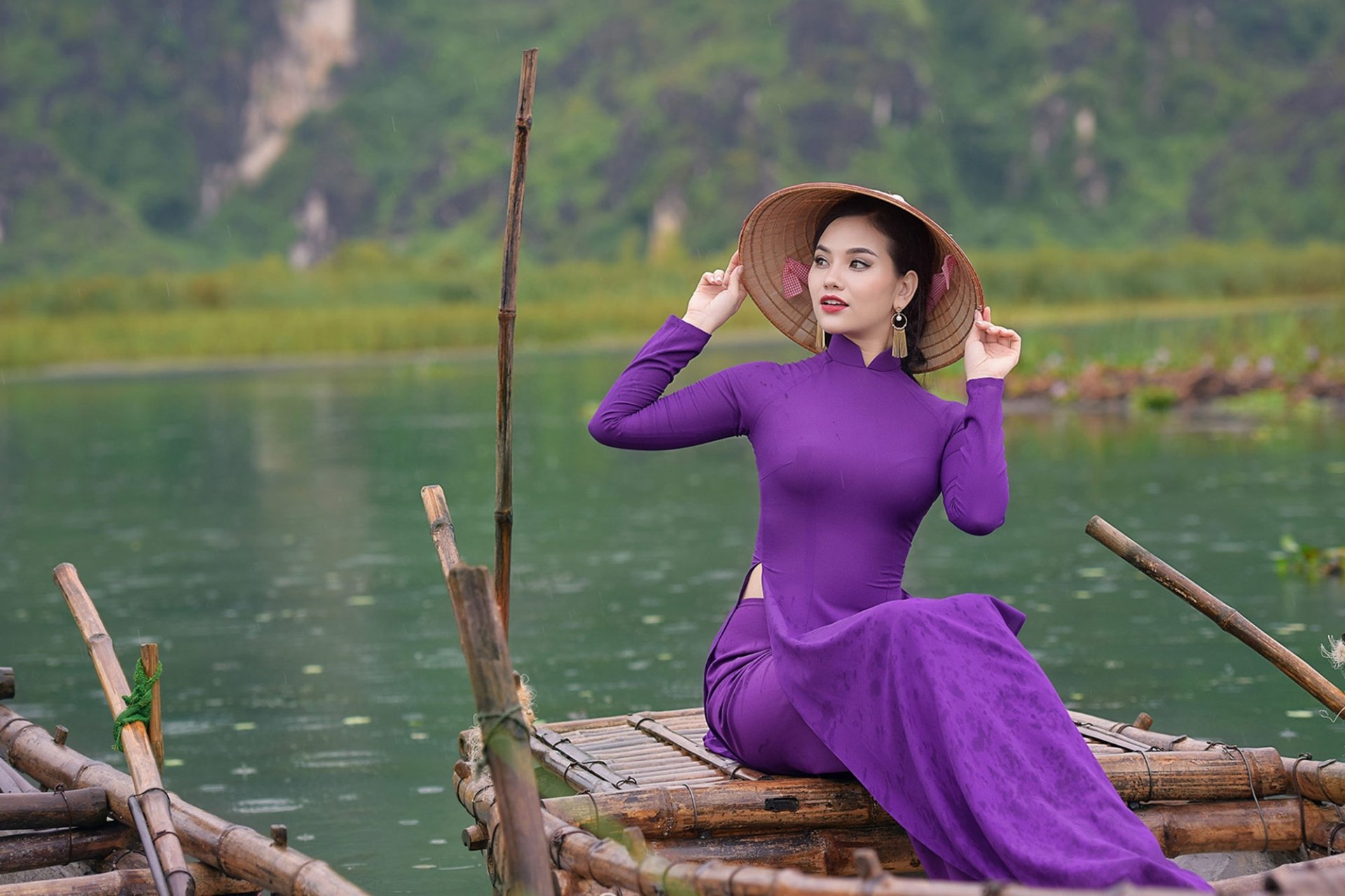 A woman wearing a purple dress and an Asian conical hat sits on a wooden raft, with a blurred green landscape in the background, captured with depth of field in this HD wallpaper.