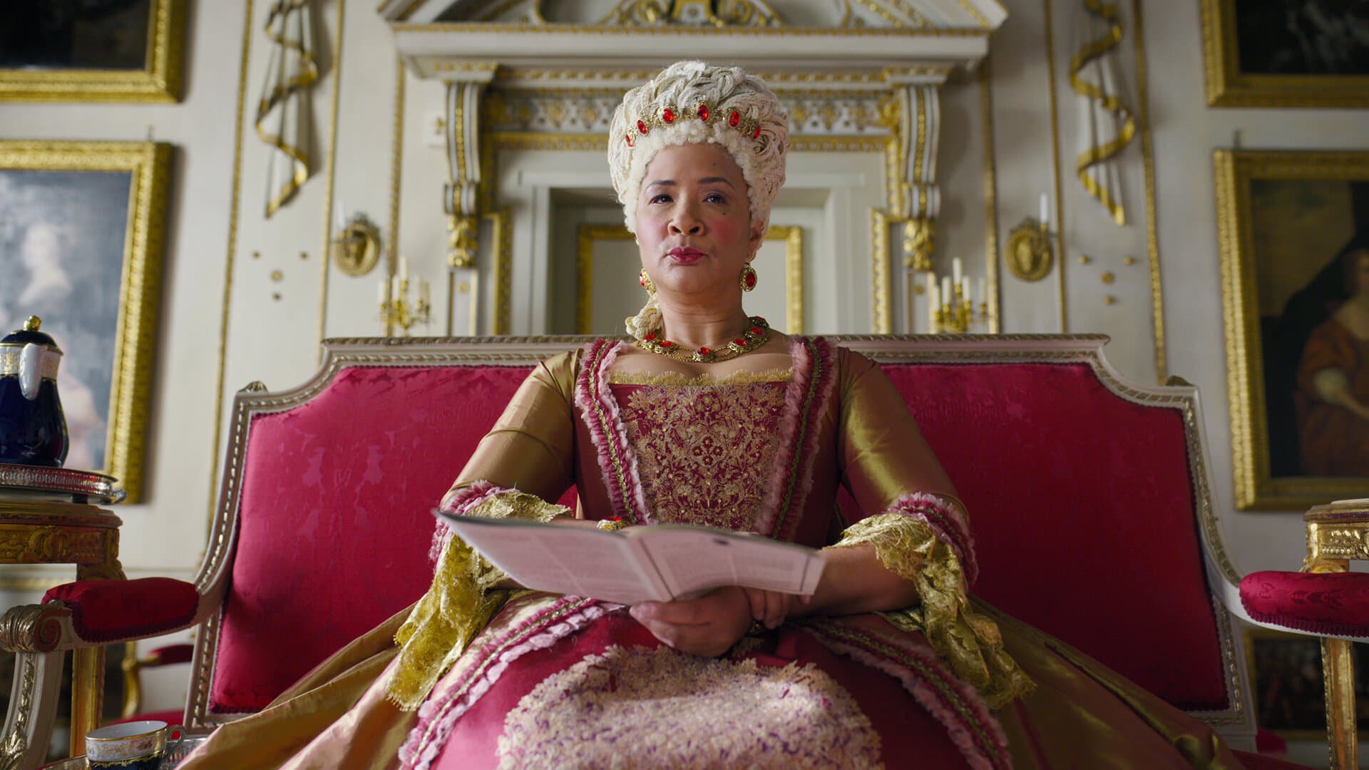 HD desktop wallpaper from Bridgerton featuring a character dressed in regal attire, sitting on a red throne in an ornate room.