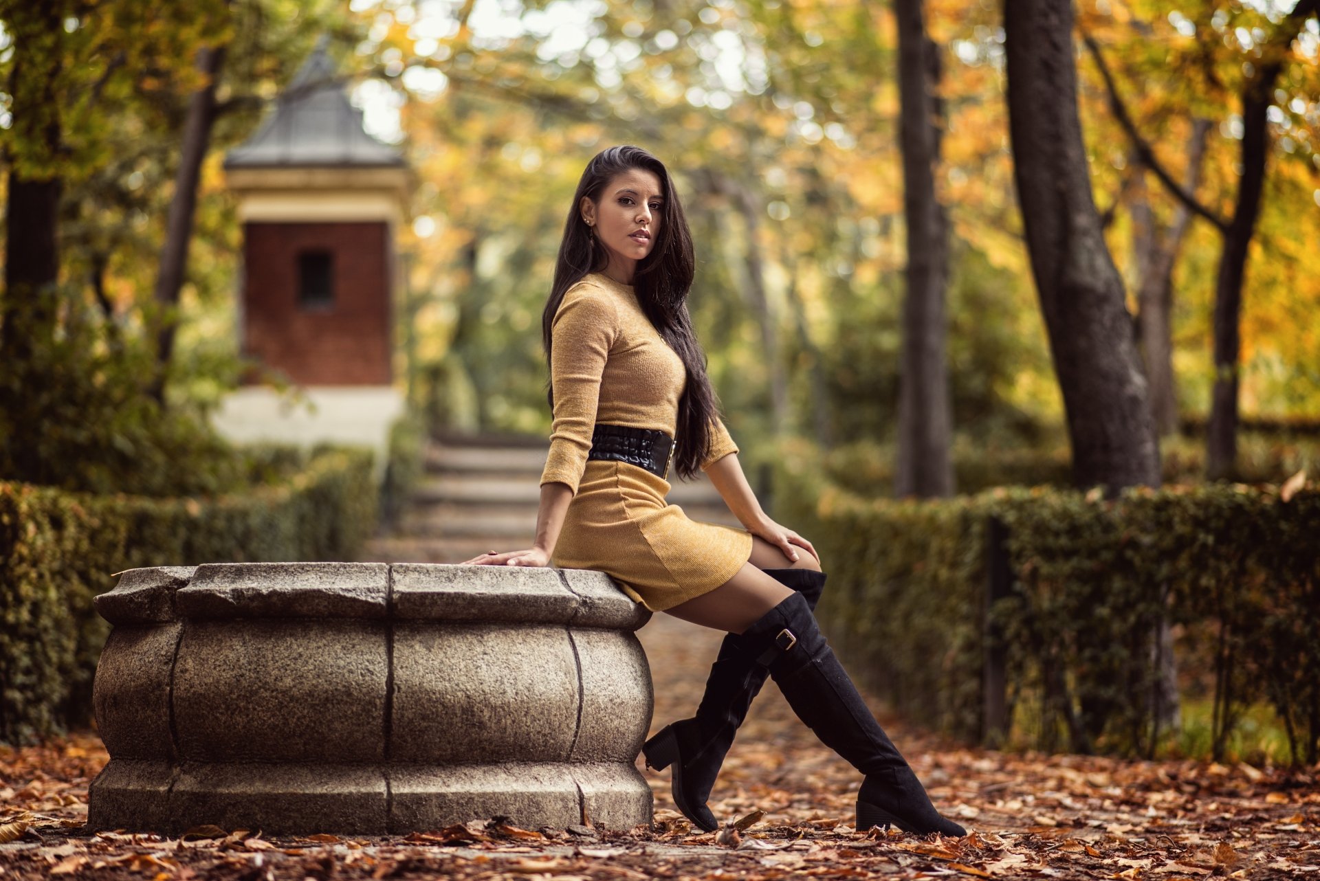 A 4K Ultra HD desktop wallpaper featuring a long-haired woman with black hair in boots, sitting on a stone structure surrounded by autumn trees, captured with depth of field.