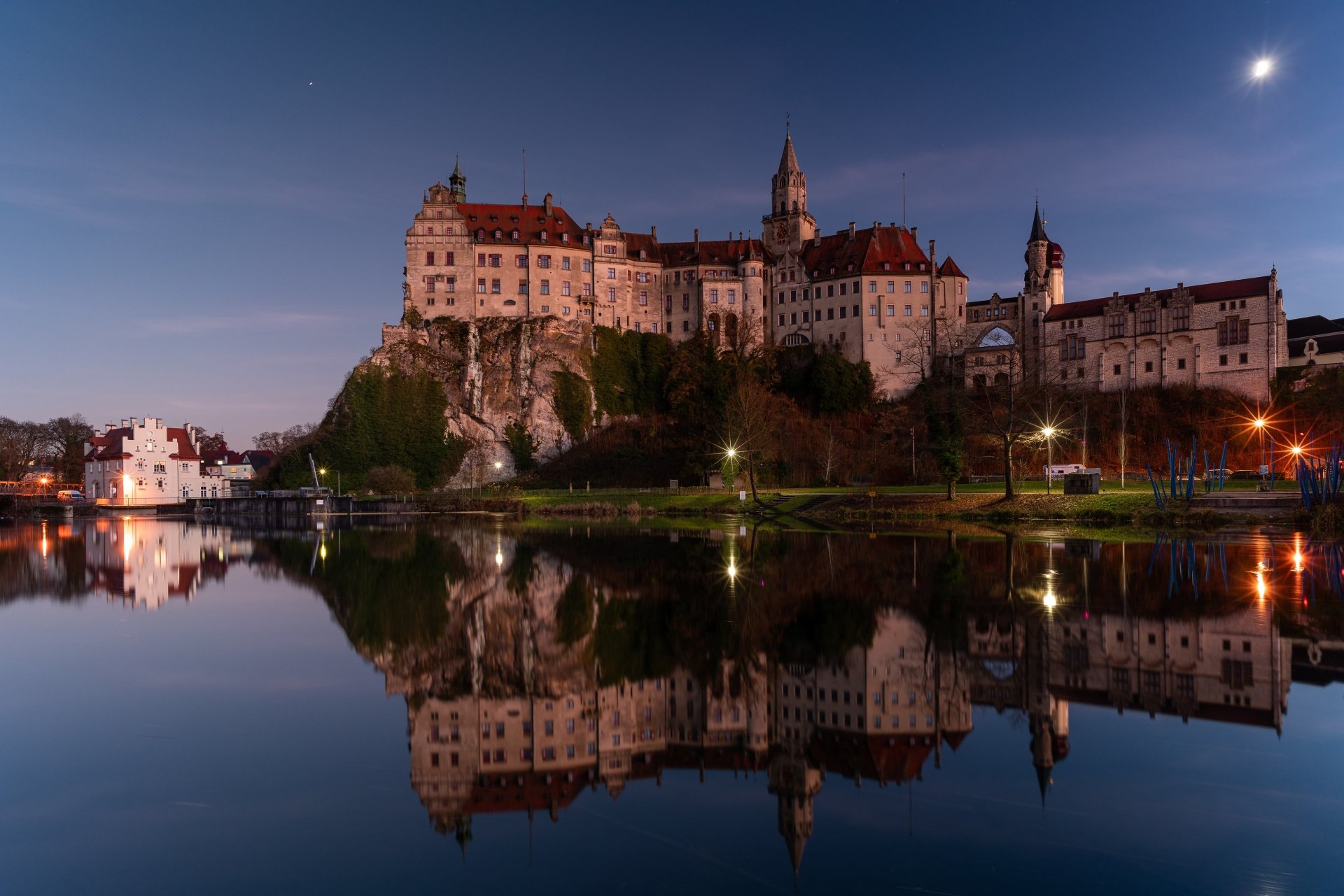 Sigmaringen Castle in Germany is beautifully illuminated at dusk, reflected clearly in the calm waters below, captured in stunning 4K Ultra HD quality.