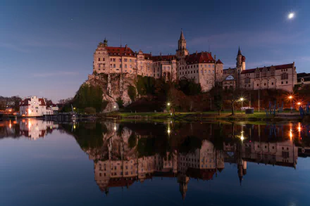 Sigmaringen Castle in Germany is beautifully illuminated at dusk, reflected clearly in the calm waters below, captured in stunning 4K Ultra HD quality.