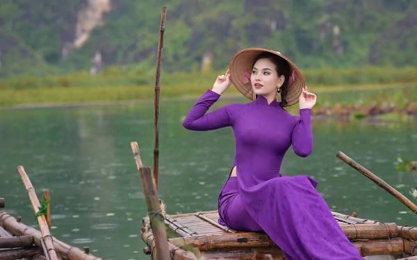 A woman wearing a purple dress and an Asian conical hat sits on a wooden raft, with a blurred green landscape in the background, captured with depth of field in this HD wallpaper.