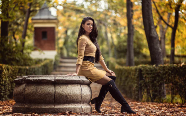 A 4K Ultra HD desktop wallpaper featuring a long-haired woman with black hair in boots, sitting on a stone structure surrounded by autumn trees, captured with depth of field.
