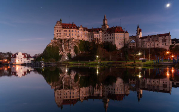 Sigmaringen Castle in Germany is beautifully illuminated at dusk, reflected clearly in the calm waters below, captured in stunning 4K Ultra HD quality.