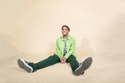 HD wallpaper featuring a smiling individual wearing a green jacket and beanie, seated with legs stretched out on a beige background.