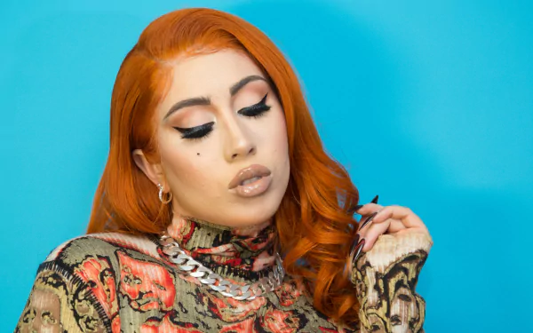 HD wallpaper featuring Kali Uchis with orange hair, bold makeup, and a printed outfit against a vibrant blue backdrop.