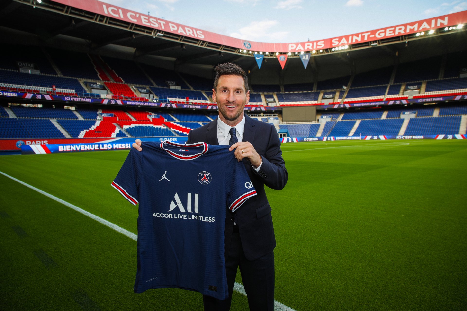 4K PC wallpaper of a footballer in a suit holding a Paris Saint-Germain jersey on the Parc des Princes pitch, with stadium stands and banners in the background.