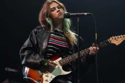 A high-definition wallpaper featuring Clairo performing with an electric guitar, exuding a cool vibe in a stylish outfit against a moody stage backdrop.