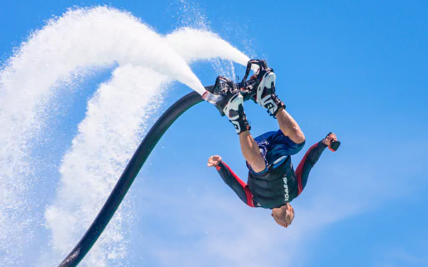HD wallpaper of a person performing an acrobatic maneuver on a flyboard against a clear blue sky.