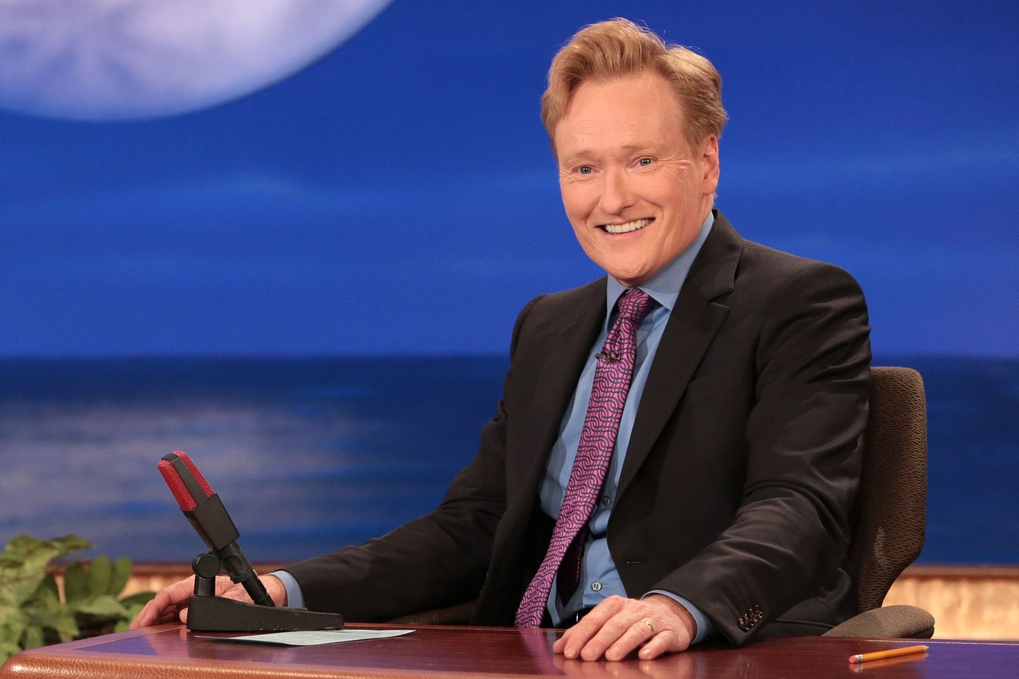 HD desktop wallpaper featuring a smiling talk show host at his desk with a moon backdrop, ideal for Conan fans.