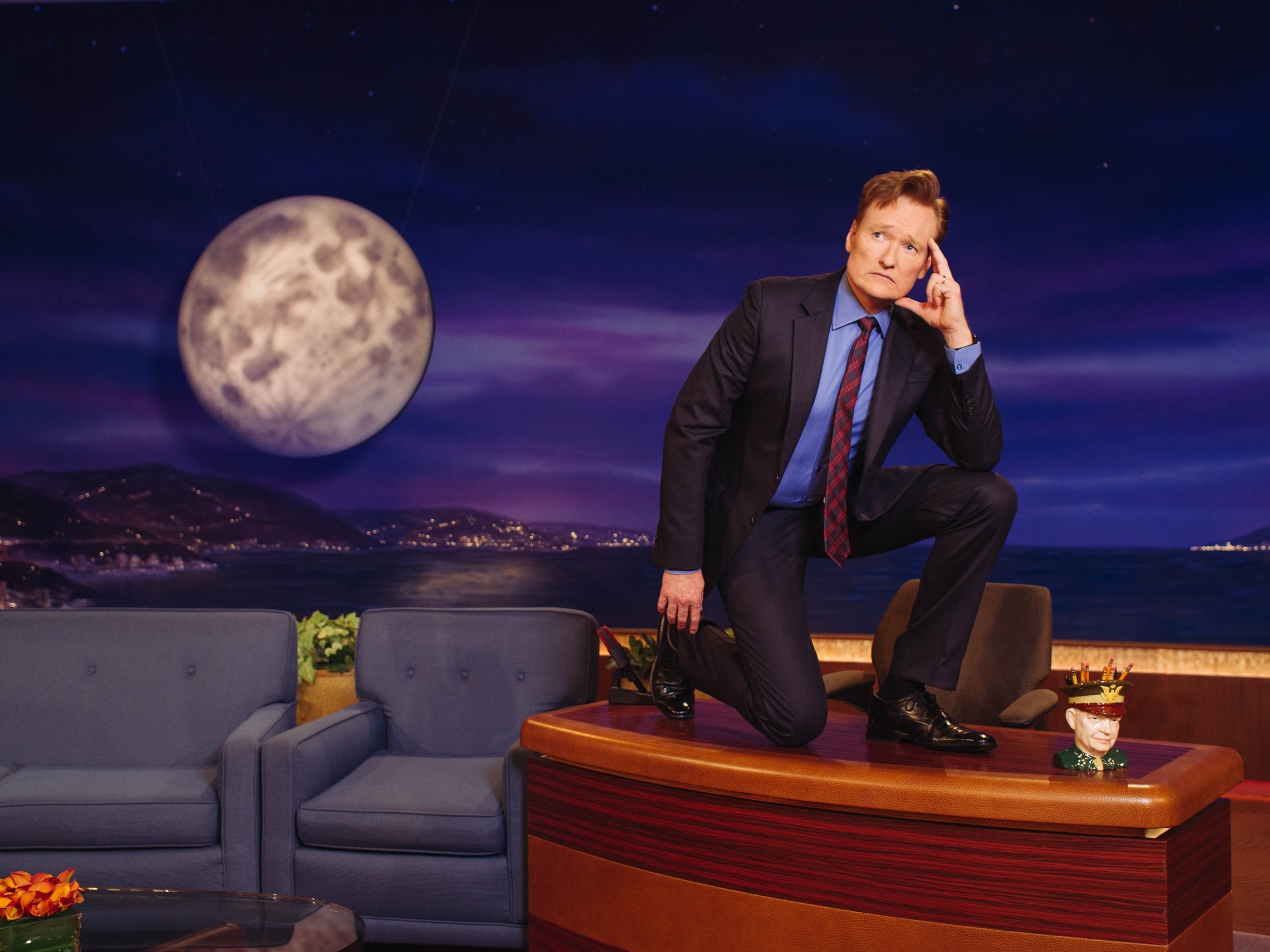 High-definition desktop wallpaper featuring a television show host posing humorously on the set with a moon backdrop and night skyline.