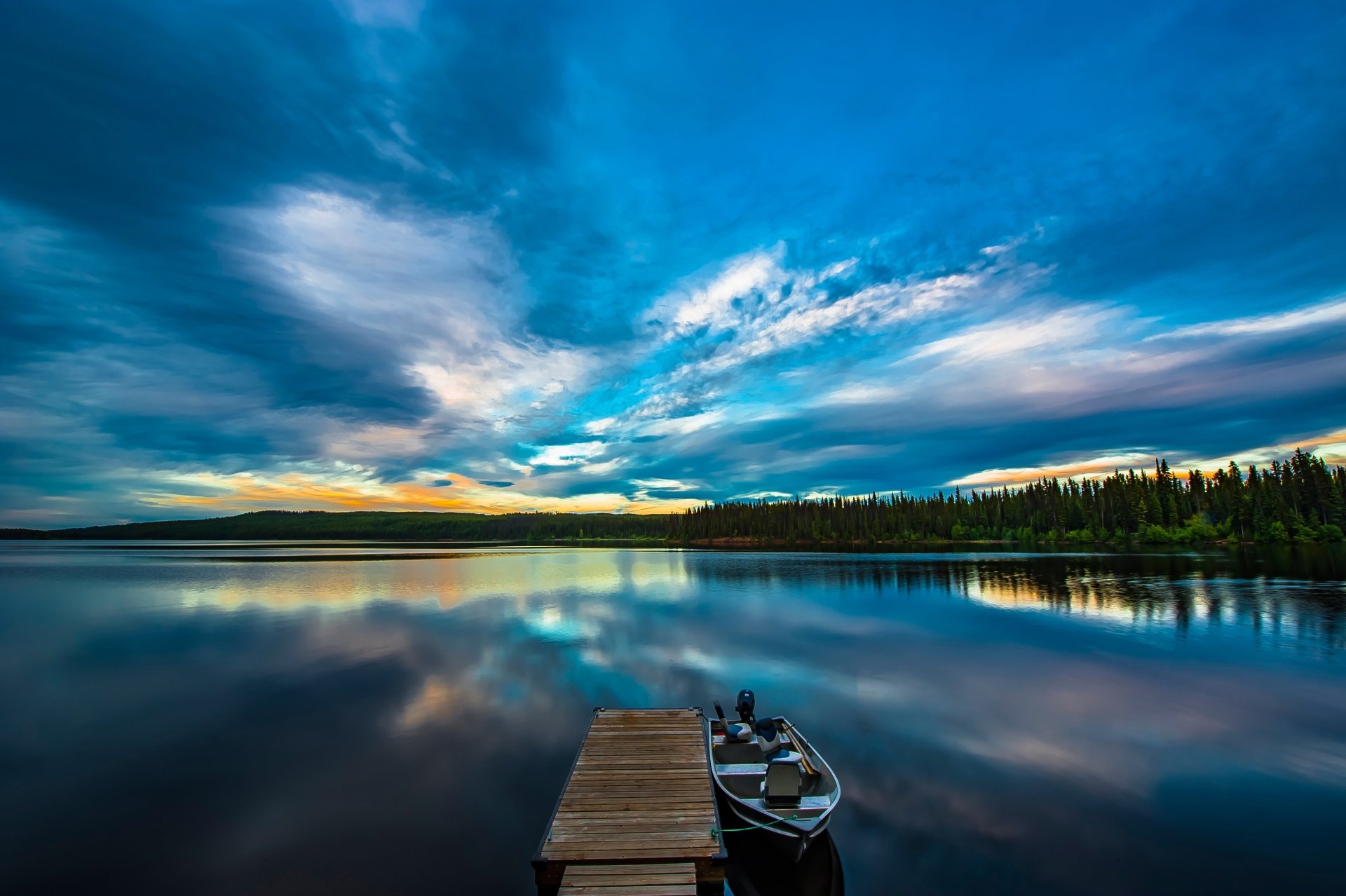 4K Ultra HD lake photography: wooden dock with a rowboat on a glassy reflective lake beneath dramatic blue sunset clouds and a distant treeline — PC desktop wallpaper/background.