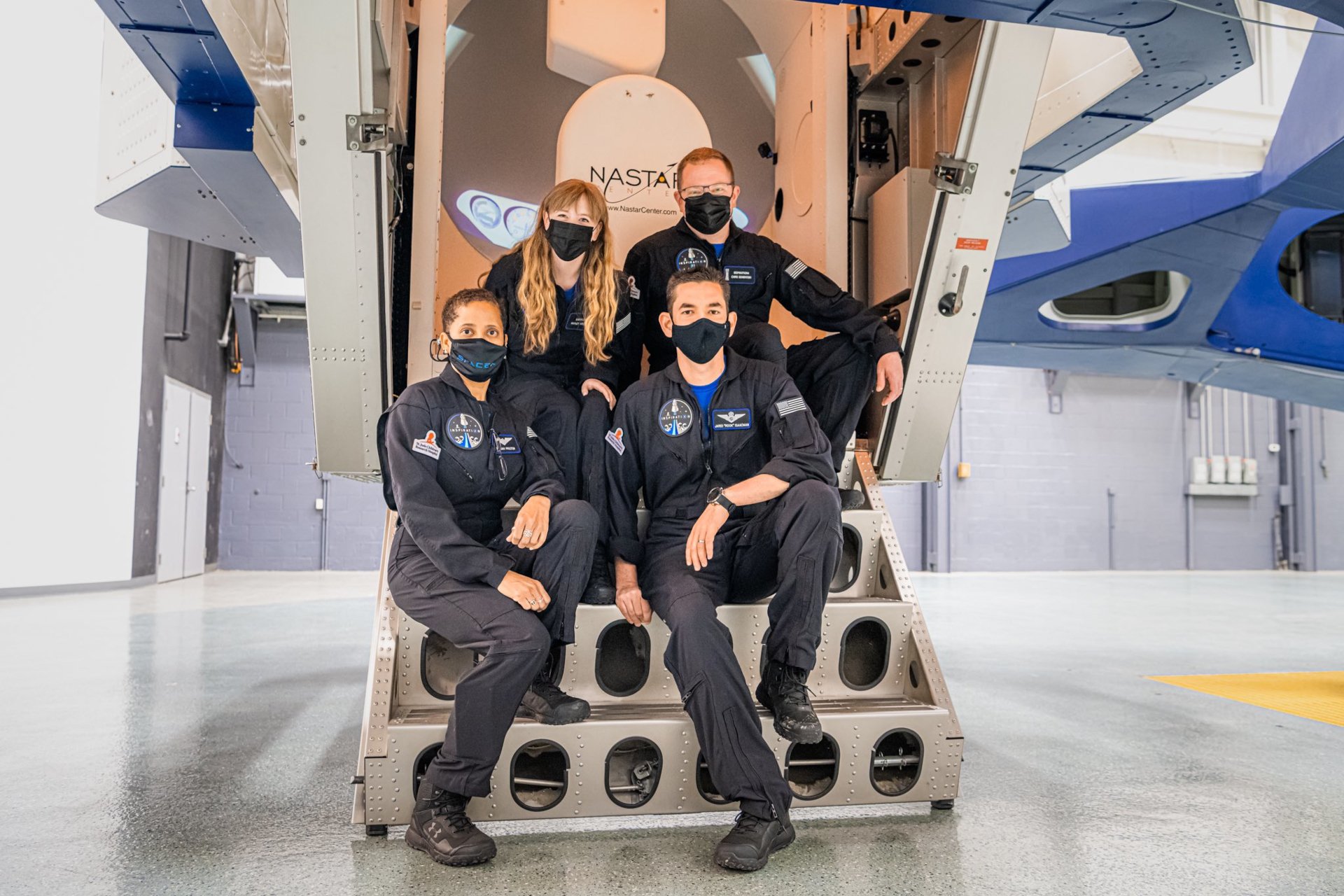 HD wallpaper of Inspiration4 crew in black flight suits and masks sitting by spacecraft hatch.
