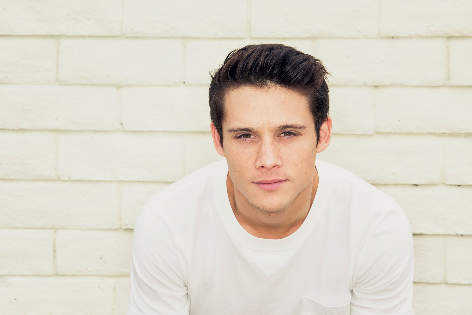 HD wallpaper featuring a portrait of a young man with dark hair, wearing a white T-shirt, set against a white brick background.