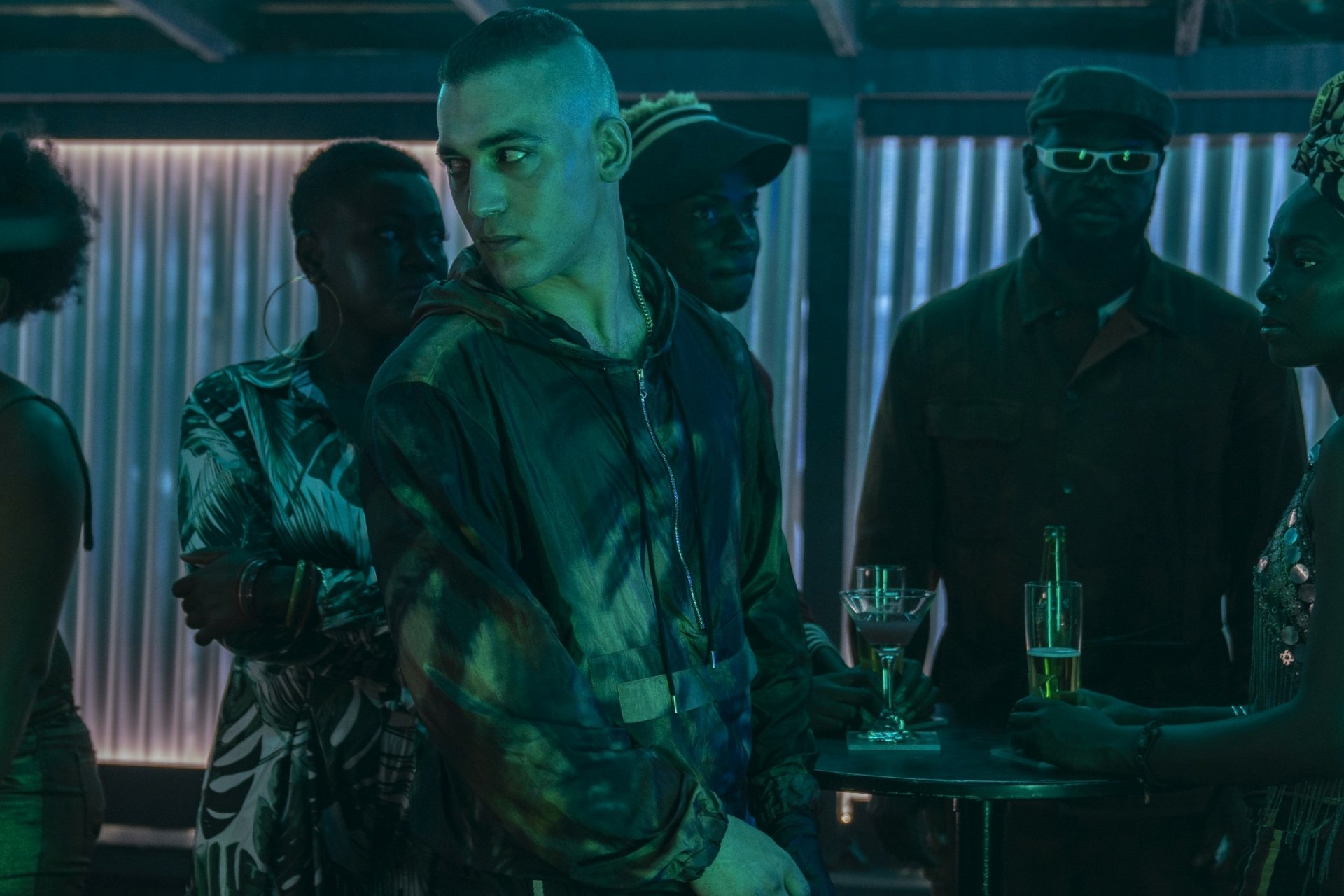 HD PC desktop wallpaper from the movie No Time to Die: a hooded man bathed in green light stands in a dim club, flanked by shadowed patrons and drinks on a small table.