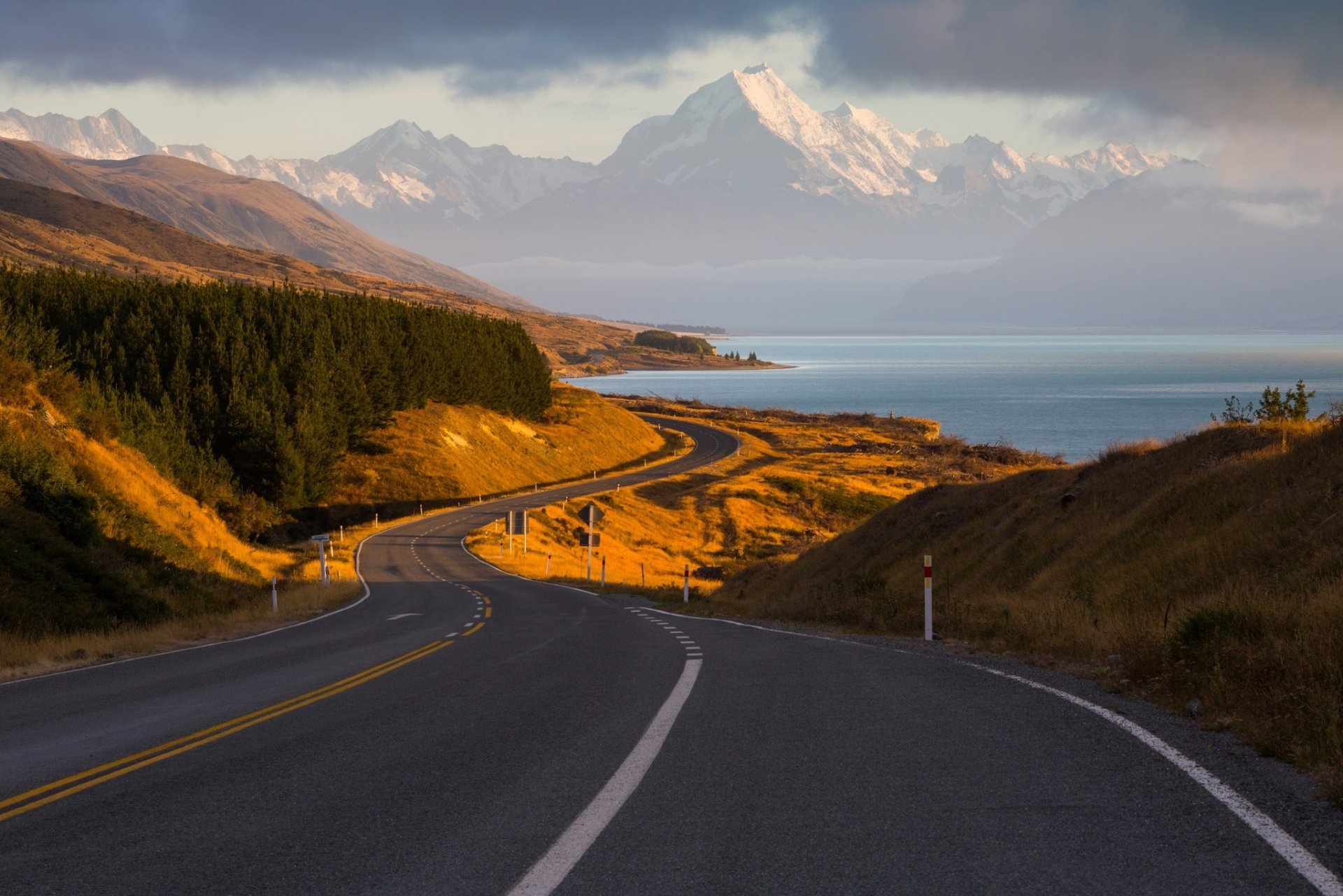 HD PC desktop wallpaper: winding man-made road through a New Zealand coastal landscape, golden hills and evergreen forest leading toward snow-capped mountains beneath a soft sky