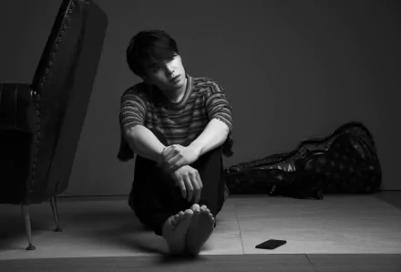 Black-and-white 4K Ultra HD wallpaper of K-pop singer Suga (Min Yoongi) from BTS sitting barefoot on the floor, with a guitar case and smartphone nearby.