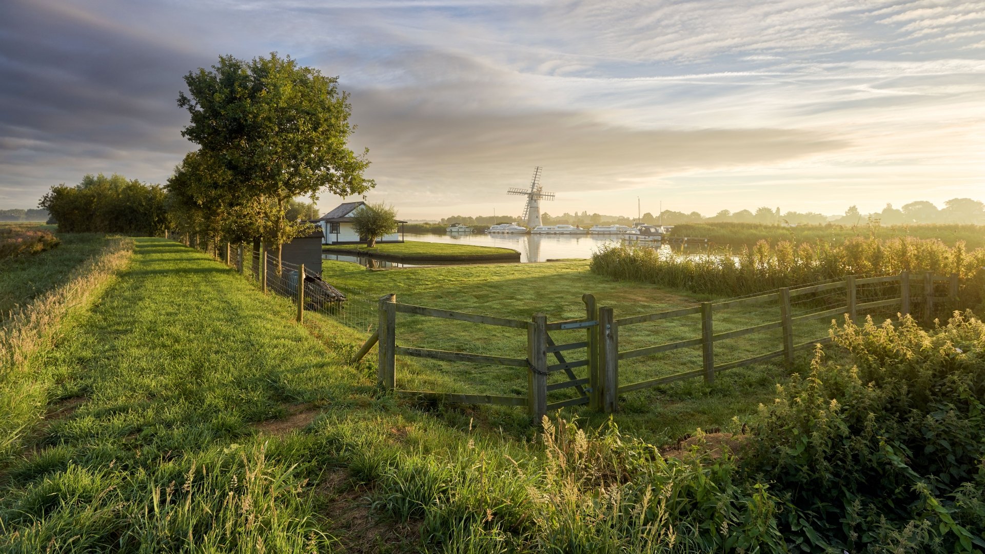 4K Ultra HD desktop wallpaper featuring a serene man-made farm landscape with lush green fields, a wooden fence, trees, and a windmill under a partly cloudy sky.