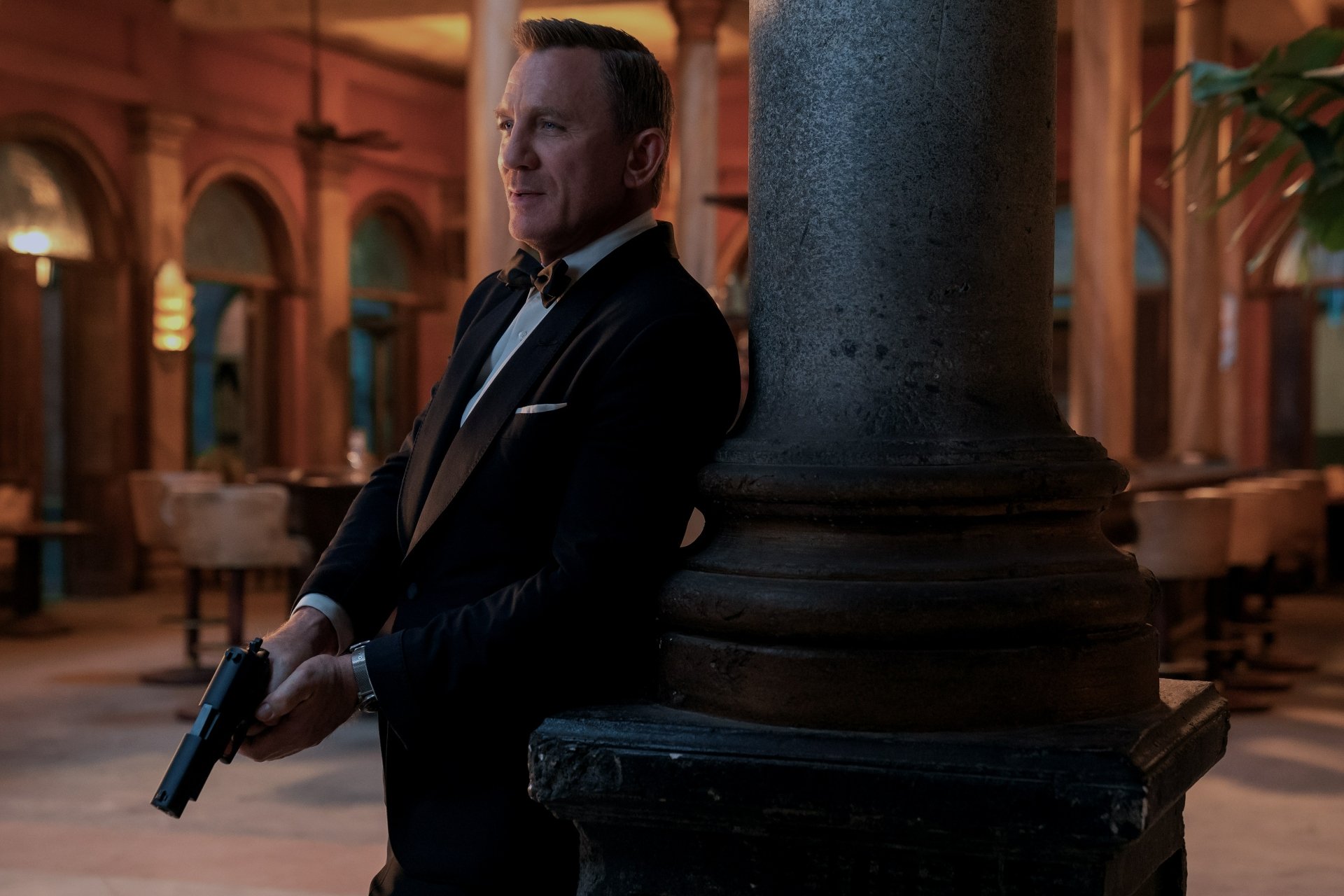 James Bond (No Time to Die) in a tuxedo, holding a pistol by a column in an opulent hall — cinematic 4K Ultra HD PC desktop wallpaper.