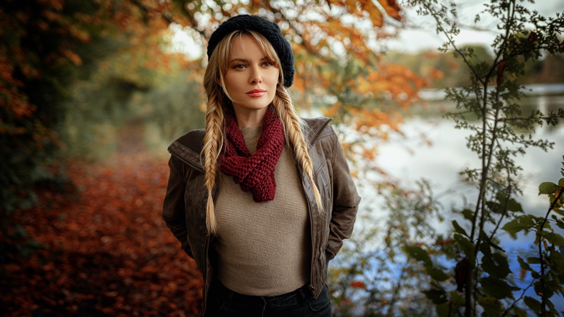 HD PC desktop wallpaper of a blonde female model with braided pigtails, wearing a beret and maroon knit scarf, standing by a lakeshore framed by warm autumn foliage.