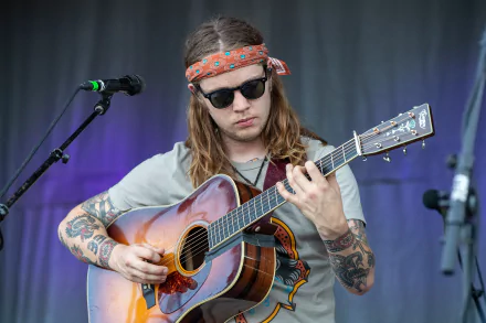 HD desktop wallpaper featuring a musician with long hair and tattoos playing an acoustic guitar on stage, with a microphone stand nearby, representing an energetic live music performance.