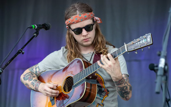 HD desktop wallpaper featuring a musician with long hair and tattoos playing an acoustic guitar on stage, with a microphone stand nearby, representing an energetic live music performance.