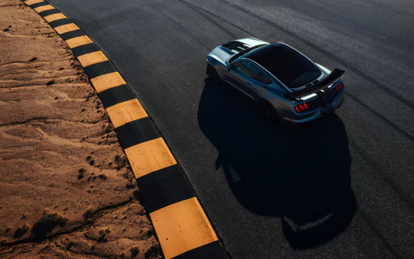 High-angle 4K desktop wallpaper of a silver Ford Mustang Shelby GT500 muscle car on a racetrack turn, casting a long shadow beside bold yellow-black curbing under dramatic lighting