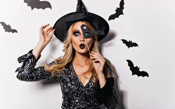 5K Ultra HD PC desktop wallpaper/background — blonde model in a witch hat and sparkly dress, holding a half-mask over one eye with bats on a white background, Halloween vibe.