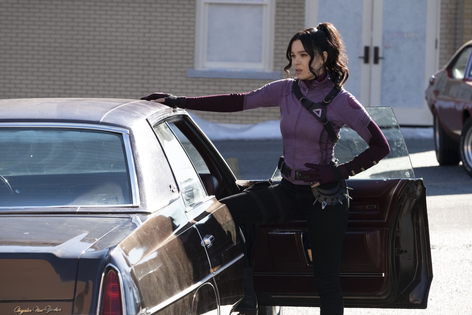 4K desktop wallpaper of a TV-show archer in a purple tactical outfit with gloves and a quiver, leaning on a car door on a sunny urban street.
