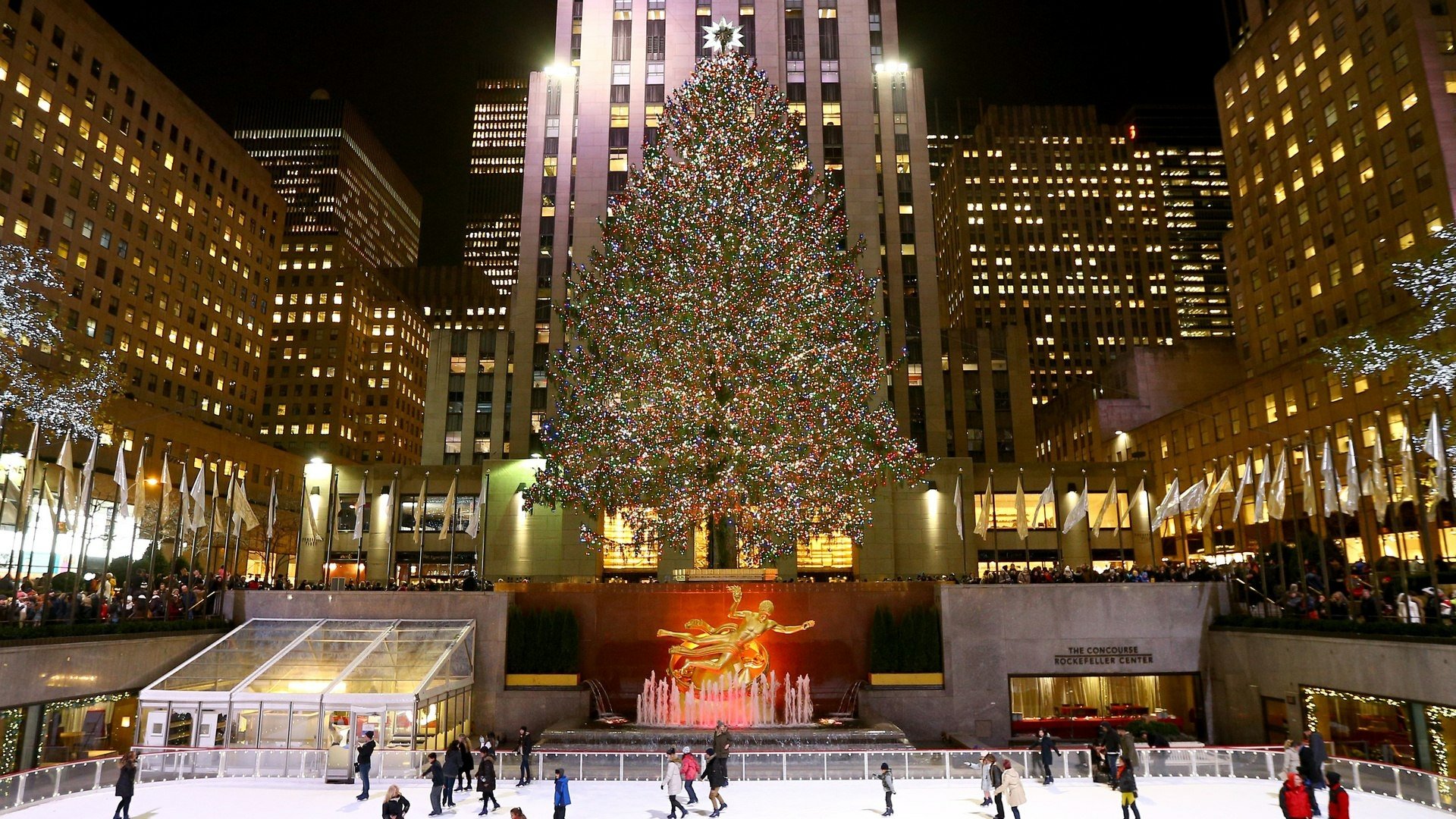 HD PC desktop wallpaper featuring a brightly lit Christmas tree at Rockefeller Center, surrounded by city buildings and an ice skating rink, capturing the holiday spirit.