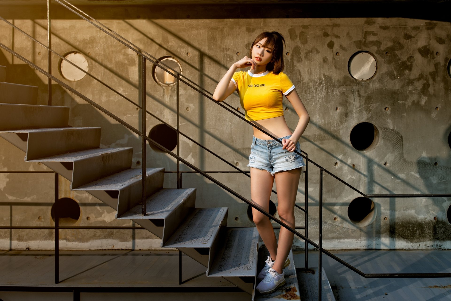 Asian woman wearing shorts and a yellow top poses on an industrial-style staircase with geometric shadows, captured in 4K Ultra HD for a PC desktop wallpaper.