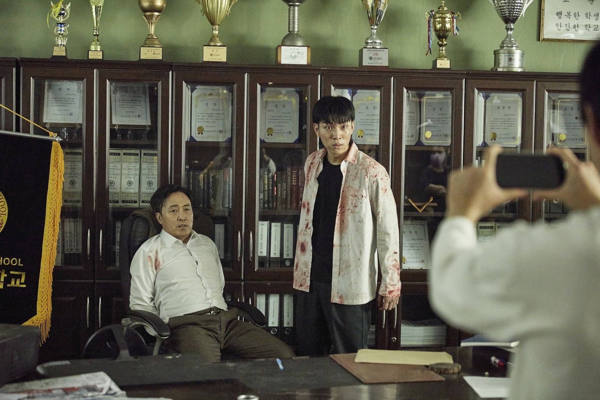 All of Us Are Dead (TV show) scene: bloodied student stands in a trophy-lined school office beside a seated man while someone photographs them — HD PC desktop wallpaper/background