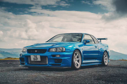 Blue Nissan Skyline R34 displayed as a high-definition wallpaper with a scenic cloudy sky background.