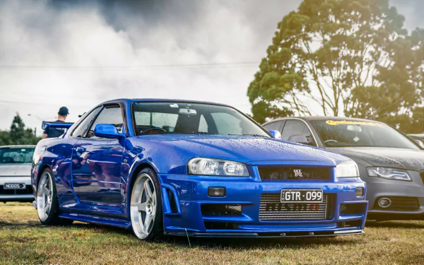 Blue Nissan Skyline R34 parked outdoors with a subtle smoky background, showcased as a high-definition desktop wallpaper.