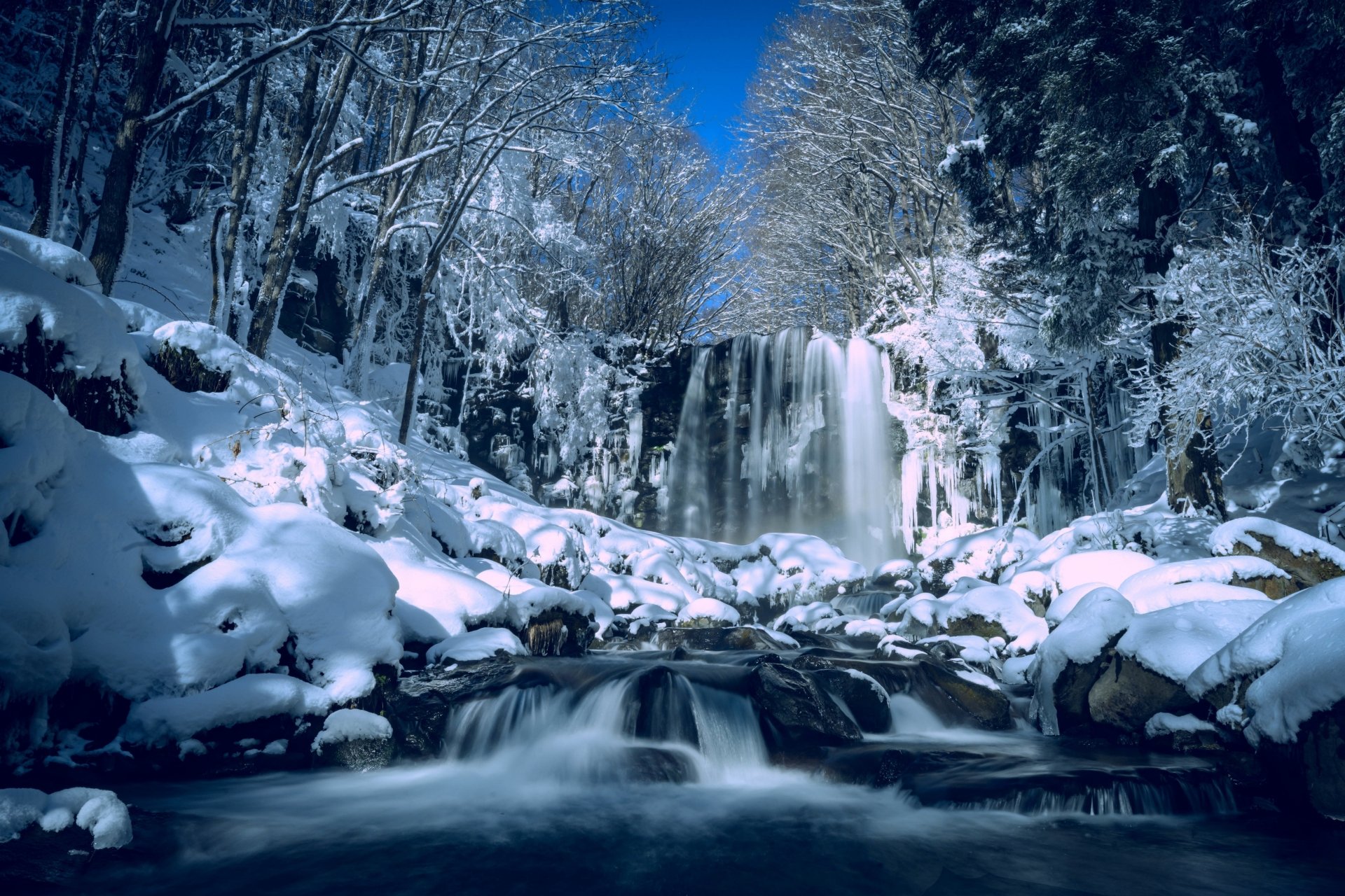 Stunning 4K Ultra HD winter landscape of a snow-covered Japanese waterfall surrounded by frosted trees under a clear blue sky.