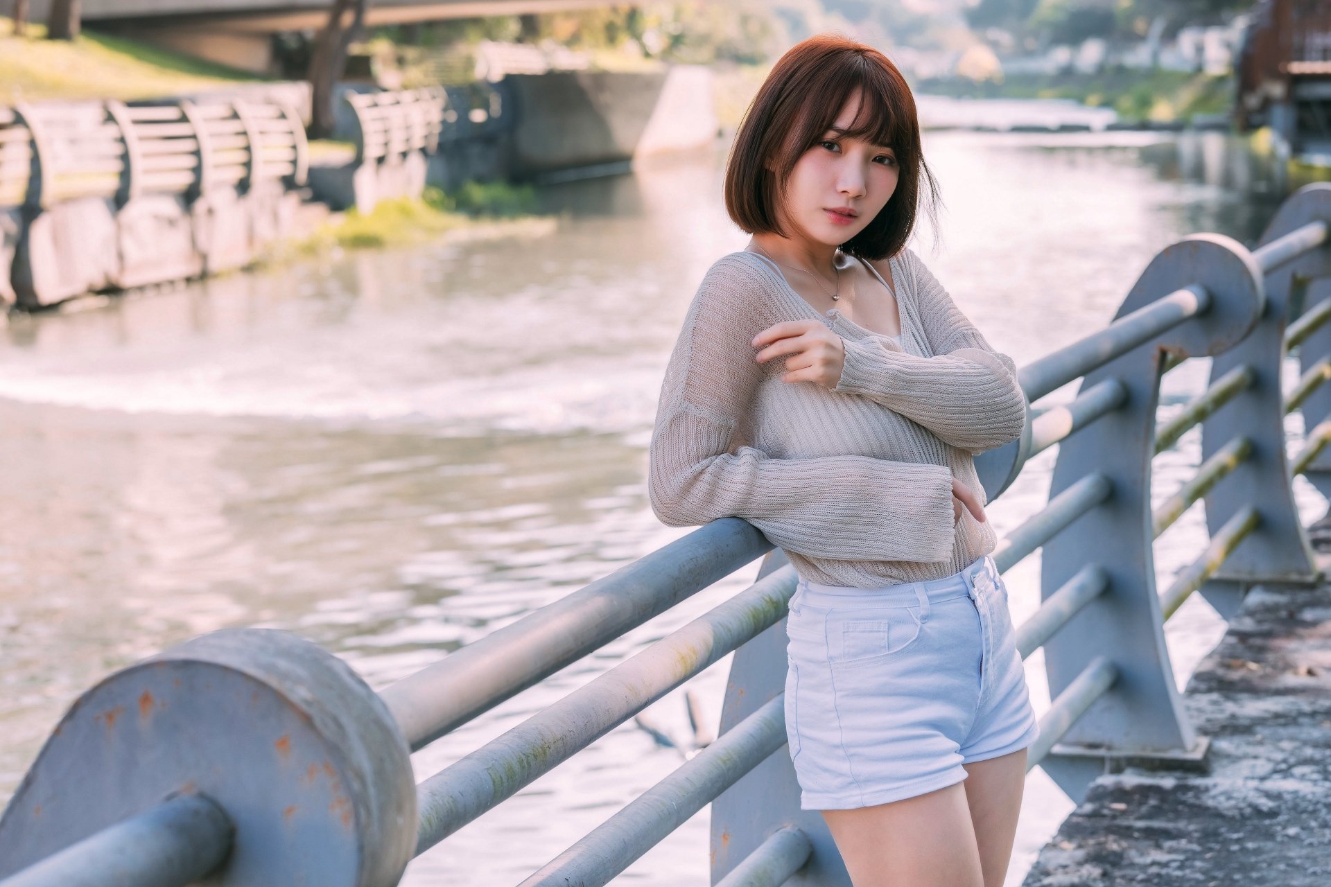 Asian woman with short hair wearing a light sweater and white shorts, leaning on a railing by a riverside, captured in high-resolution 4K Ultra HD detail.