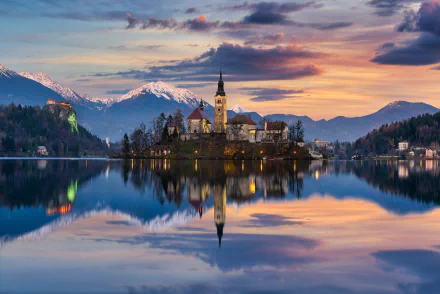 Sunset view of the Assumption of Mary Church reflected in the calm waters of Lake Bled, Slovenia, with the surrounding mountains under a colorful sky.