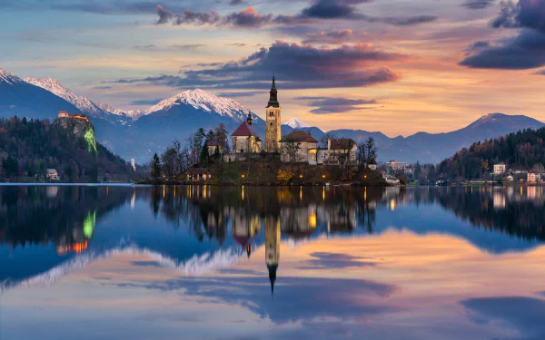 Sunset view of the Assumption of Mary Church reflected in the calm waters of Lake Bled, Slovenia, with the surrounding mountains under a colorful sky.