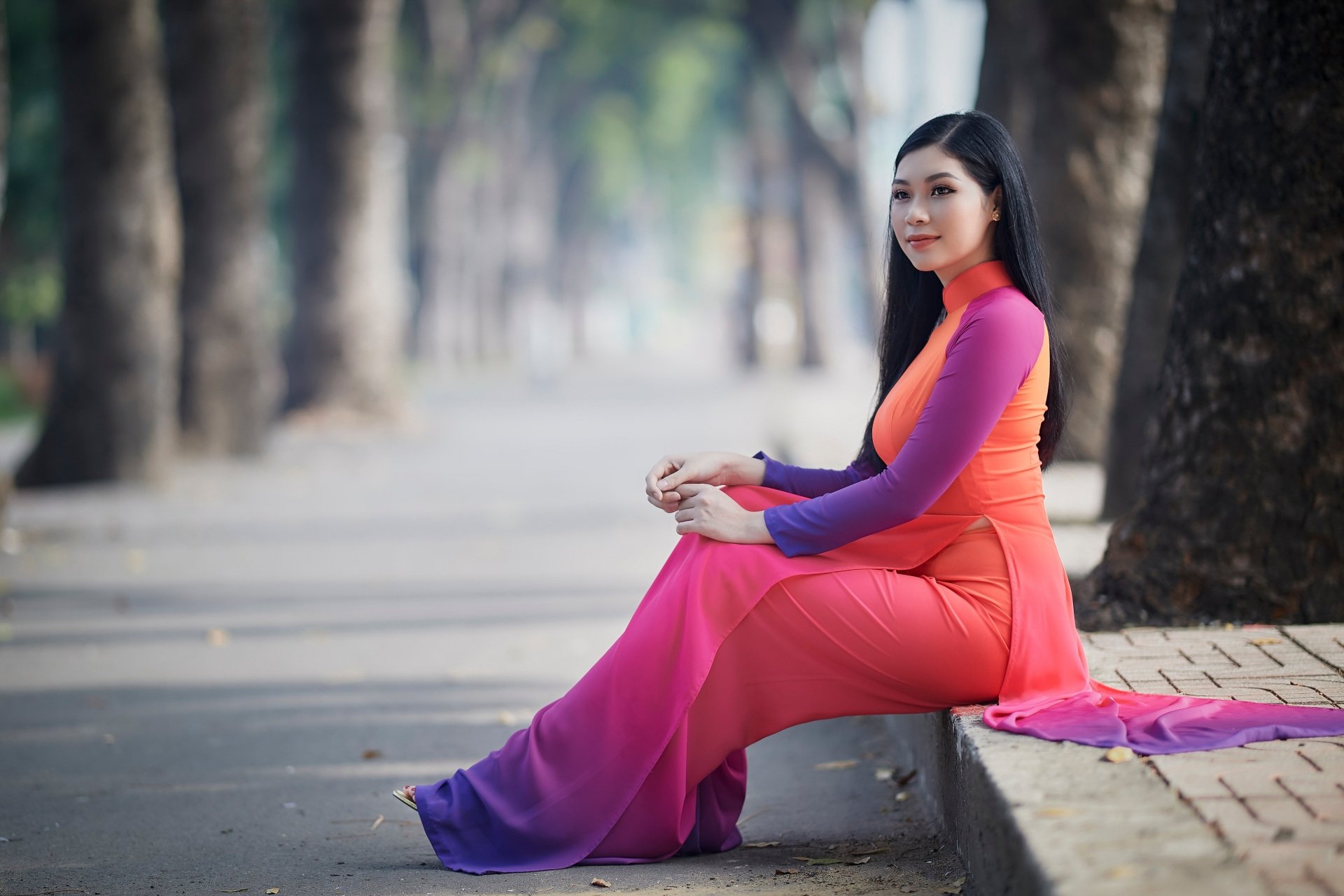 Asian woman in vibrant pink and orange traditional dress sitting on a sidewalk, captured in sharp 4K Ultra HD quality, framed by a tree-lined path background.