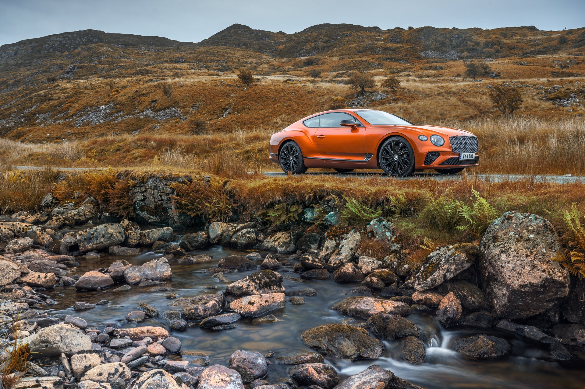 Orange Bentley Continental GT on a rugged moor beside a rocky stream under overcast sky — 5K Ultra HD PC desktop wallpaper/background.