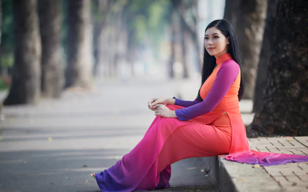 Asian woman in vibrant pink and orange traditional dress sitting on a sidewalk, captured in sharp 4K Ultra HD quality, framed by a tree-lined path background.