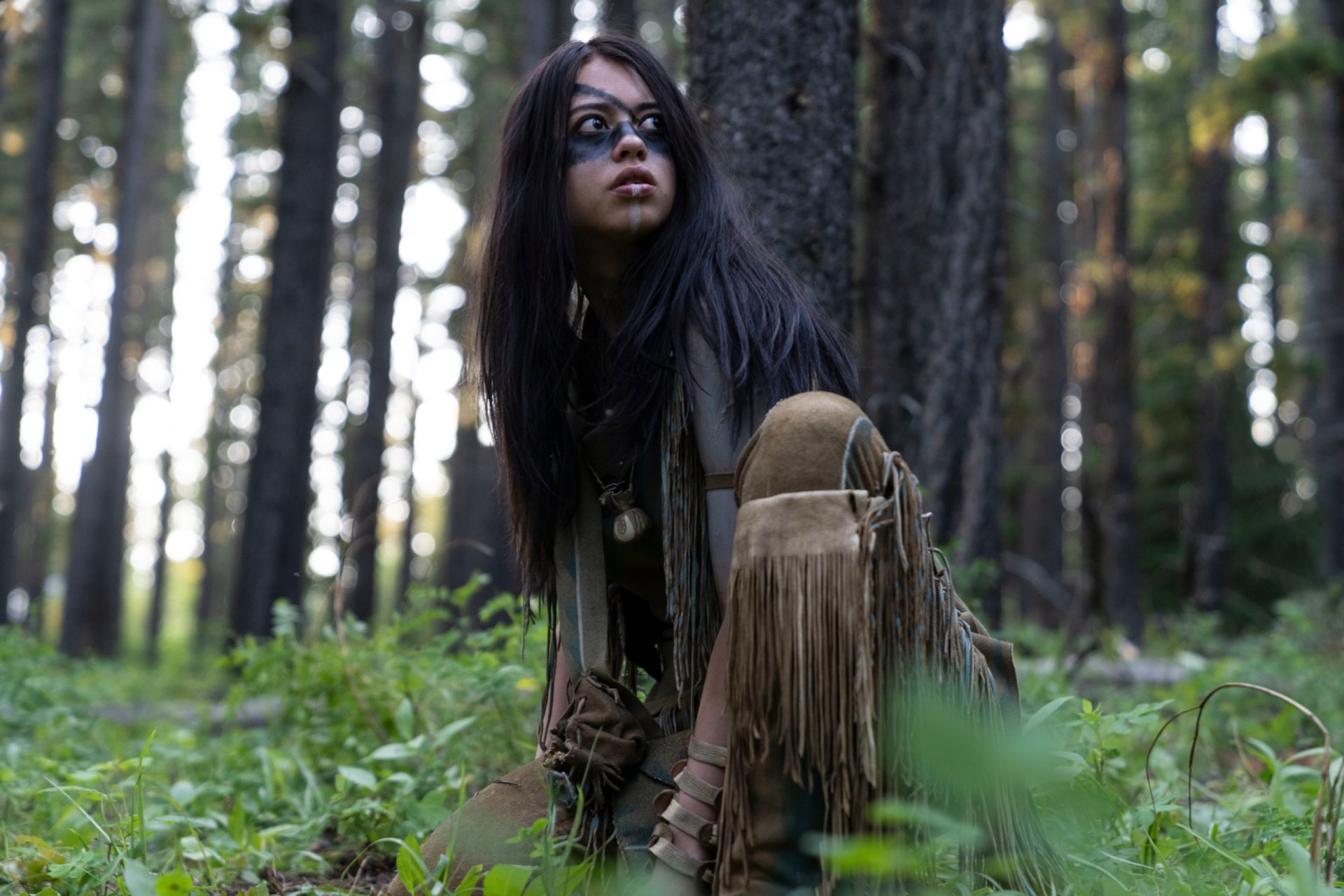 Young woman with face paint and fringed buckskin crouches in a pine forest; still from the movie Prey (2022) — 2K Quad HD PC desktop wallpaper.